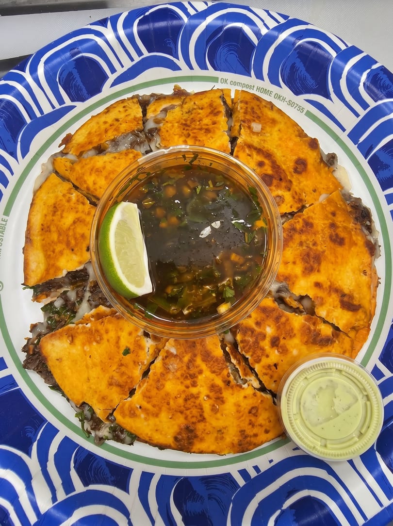 A large, golden-brown quesadilla, cut into wedges, with a side of dark dipping sauce and a lime wedge.