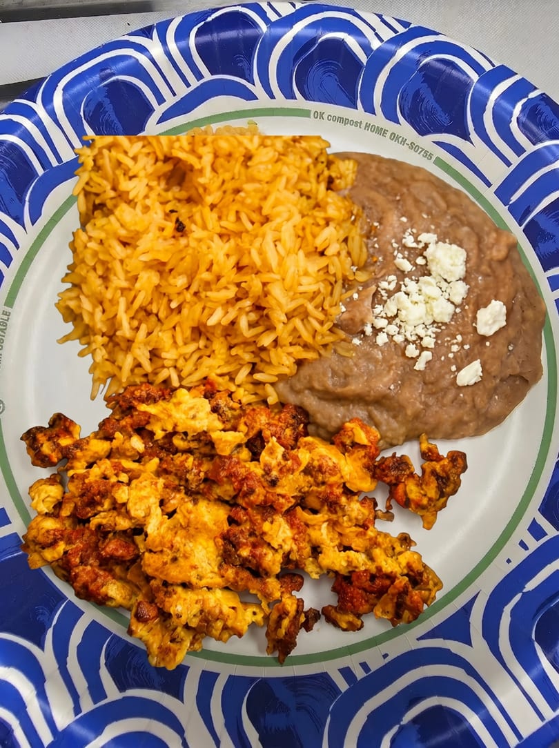 A paper plate with Mexican rice, refried beans topped with cheese, and scrambled eggs with chorizo.
