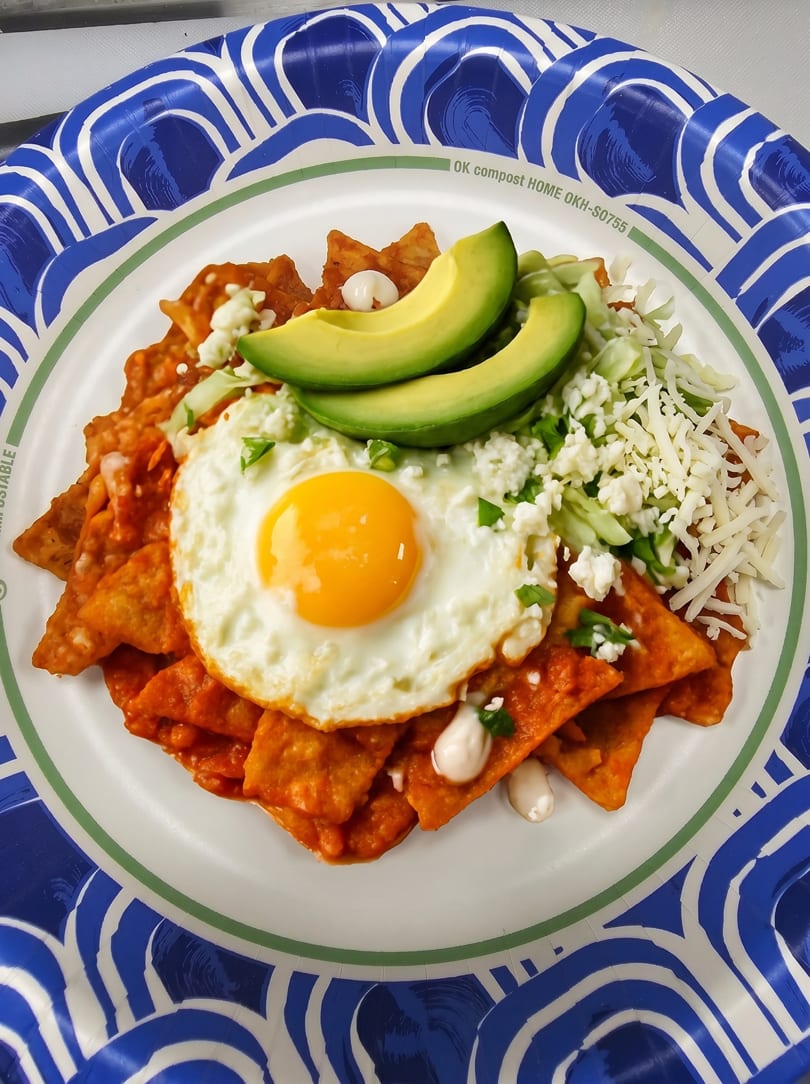 Chilaquiles with a fried egg, avocado slices, shredded cheese, and crema on a paper plate.