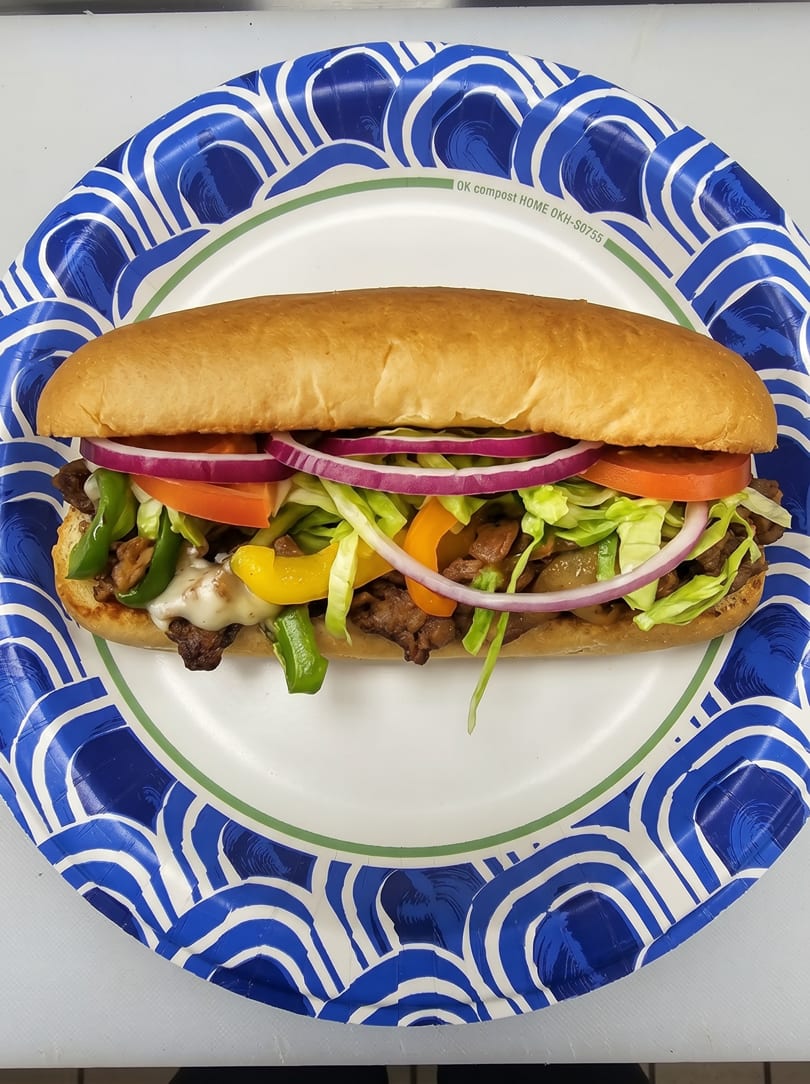 A steak and cheese sub with lettuce, tomato, onions, and peppers on a paper plate.