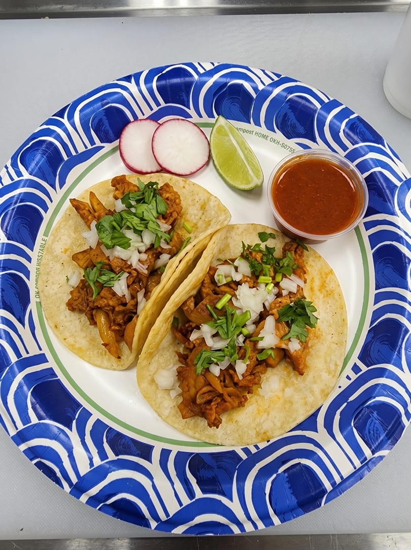 Two tacos with meat, onions, and cilantro, served with radishes, lime, and salsa on a paper plate.